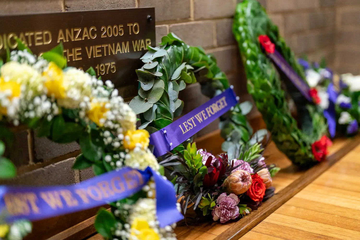 Commemorative wreaths and flowers placed beneath Vietnam War memorial plaque with “Lest We Forget” ribbons