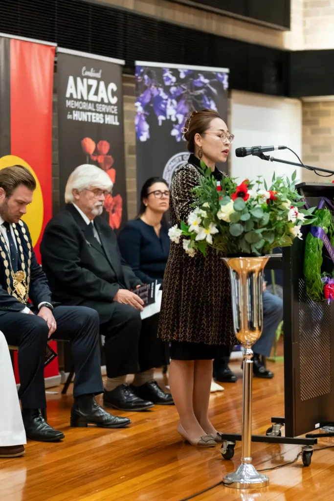 Female speaker addressing audience at ANZAC remembrance ceremony with officials seated behind
