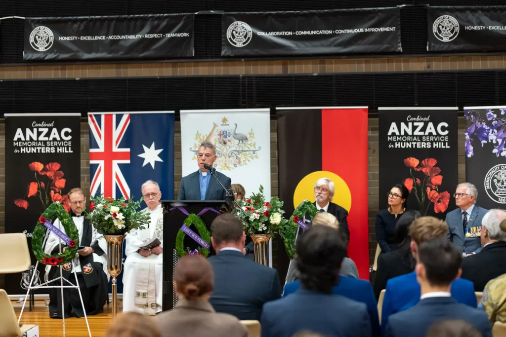 Wide interior view of ANZAC memorial service stage with speakers, banners, wreaths, and audience