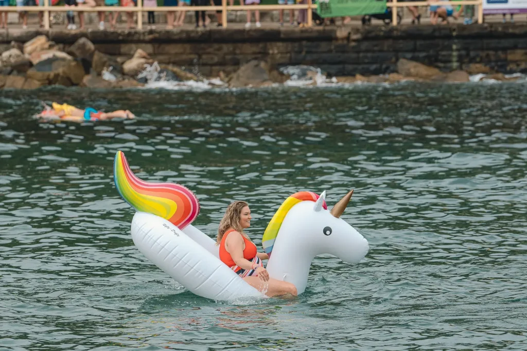 Woman riding a large white rainbow unicorn inflatable across the water at Manly